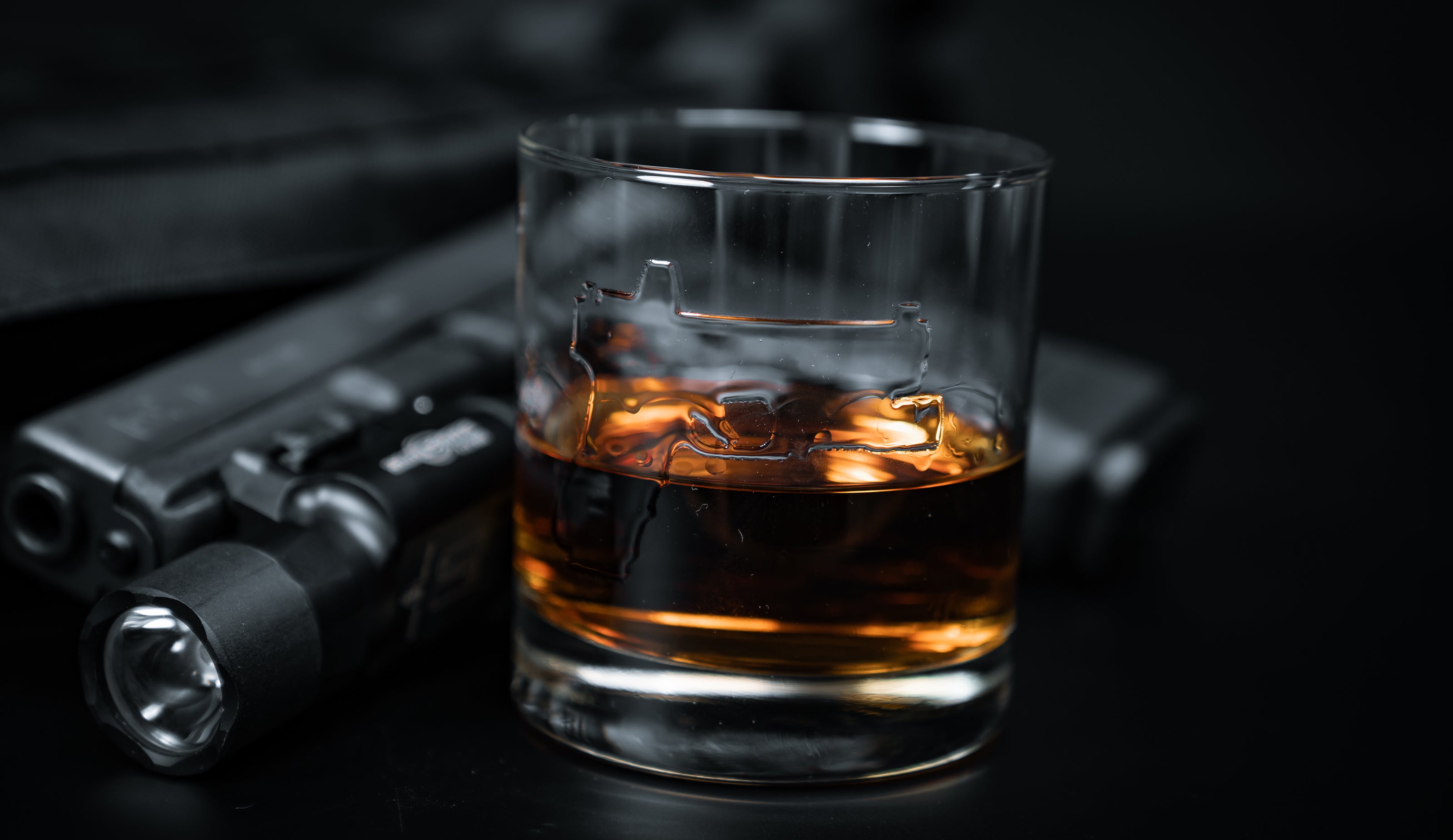 whiskey glass embossed with a handgun, half full of bourbon, in front of a handgun on a black table