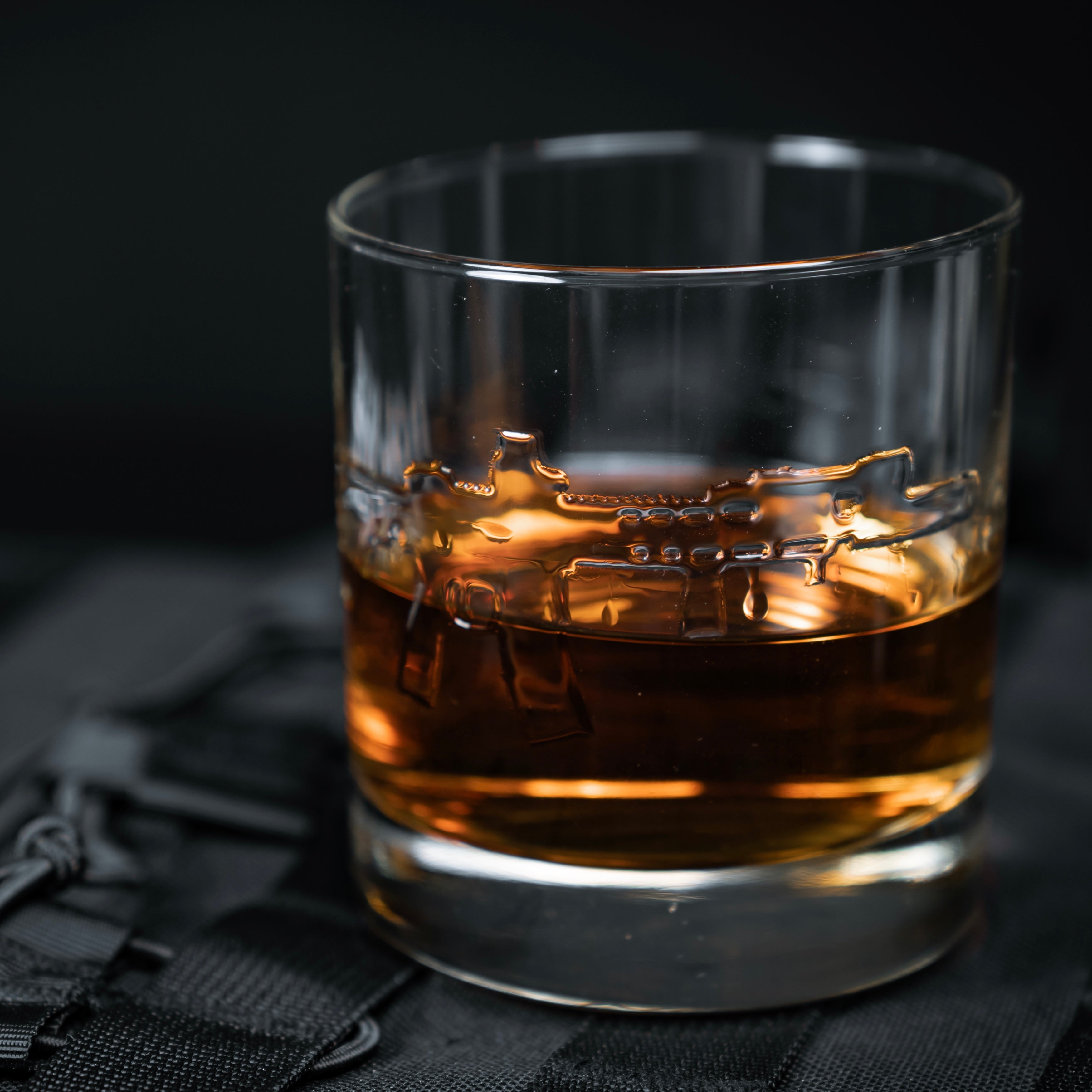 whiskey rocks glass embossed with an AR15, half full with bourbon, on a molle plate carrier