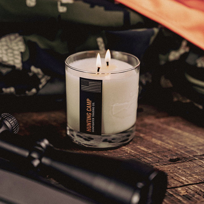 hunting camp candle on wood table, scoped rifle laying in front and camouflage clothing behind