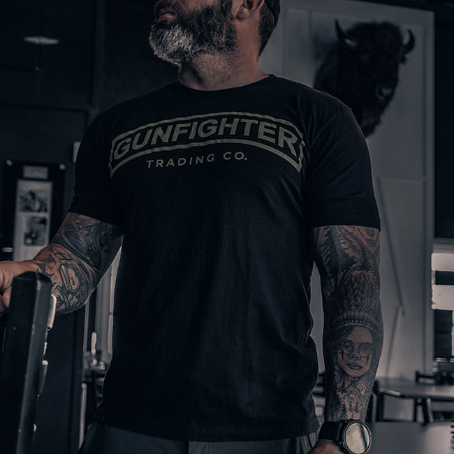 man standing wearing a black t-shirt with a printed Gunfighter Trading Co. tab logo in green